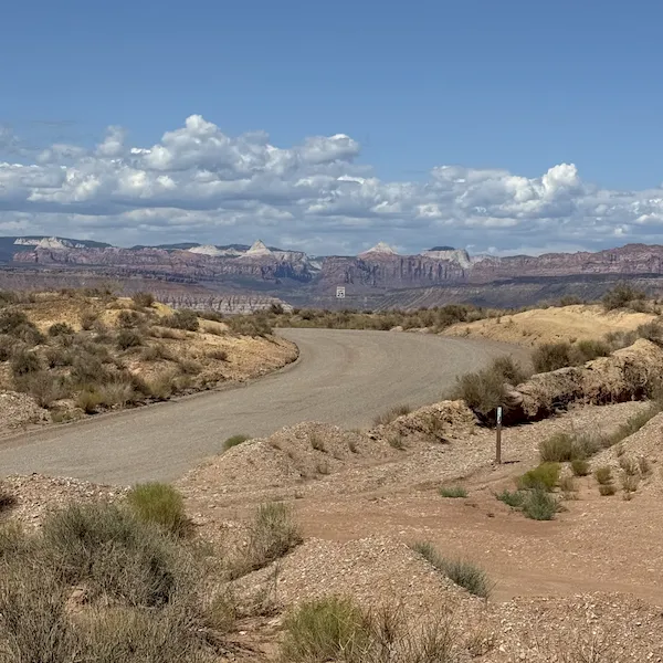 An image of a dirt road winding through a desert toward distant mountains under a cloudy sky, with a small, distant 25 mph speed limit sign in the Zion National Park area.
