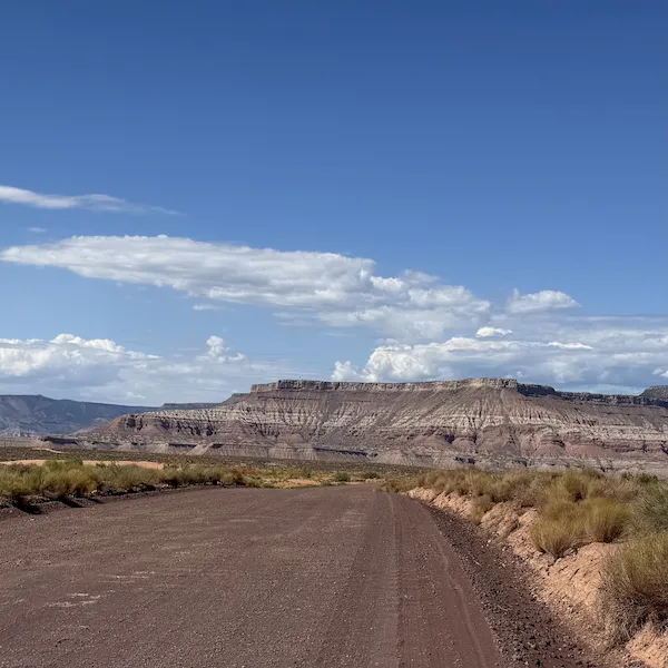 An image of a dirt road stretching through a desert landscape leading towards a mountain range under a blue sky in the Zion National Park area.