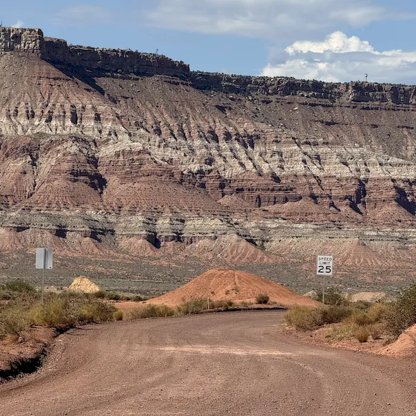 An image of a dirt road in a desert canyon leading toward red mountains and a 25 mph speed limit sign on the right side of the road  in the Zion National Park area.