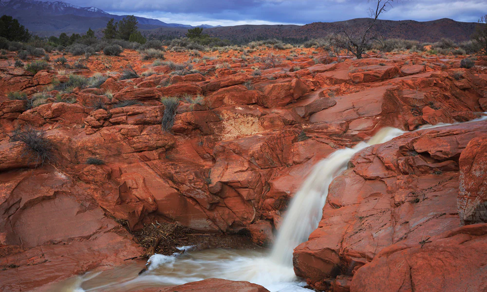 Red rock waterfall cascading over rugged terrain with desert vegetation
