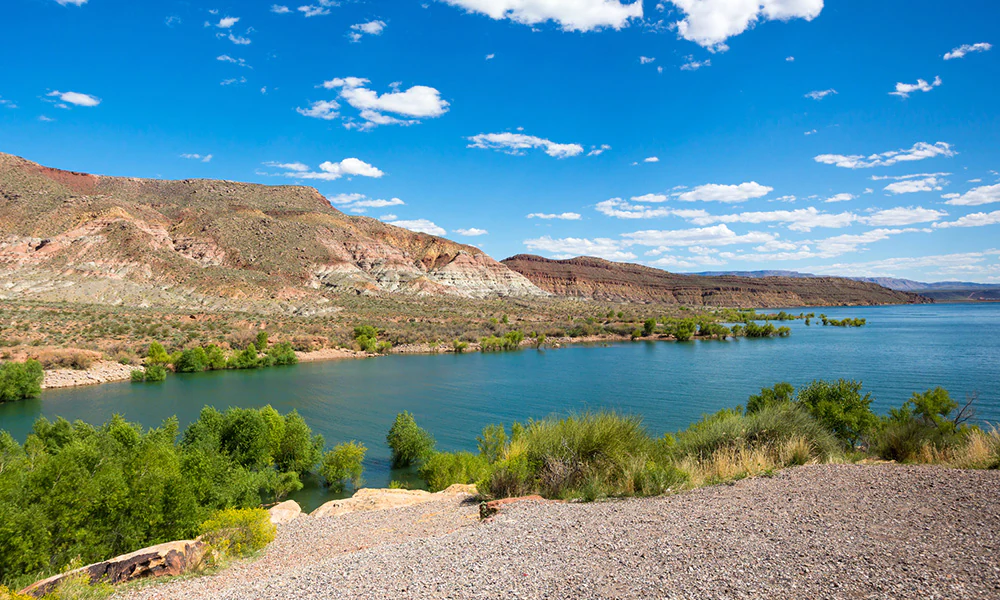 Blue lake with rocky mountains, greenery, and gravel shore under clear sky