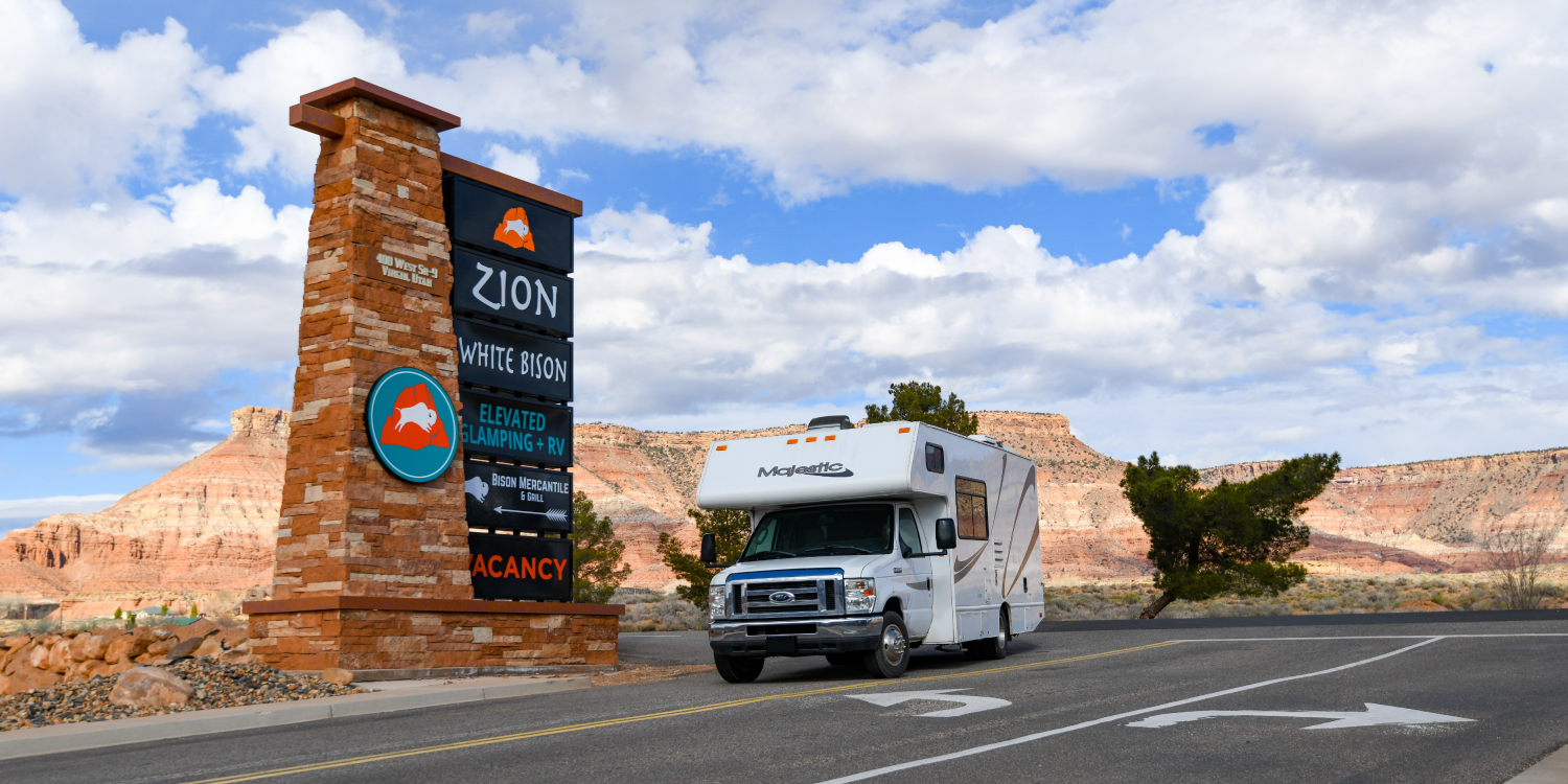 An RV turns into the driveway of Zion White Bison Resort. A stacked stone sign stands tall with the resort logos.